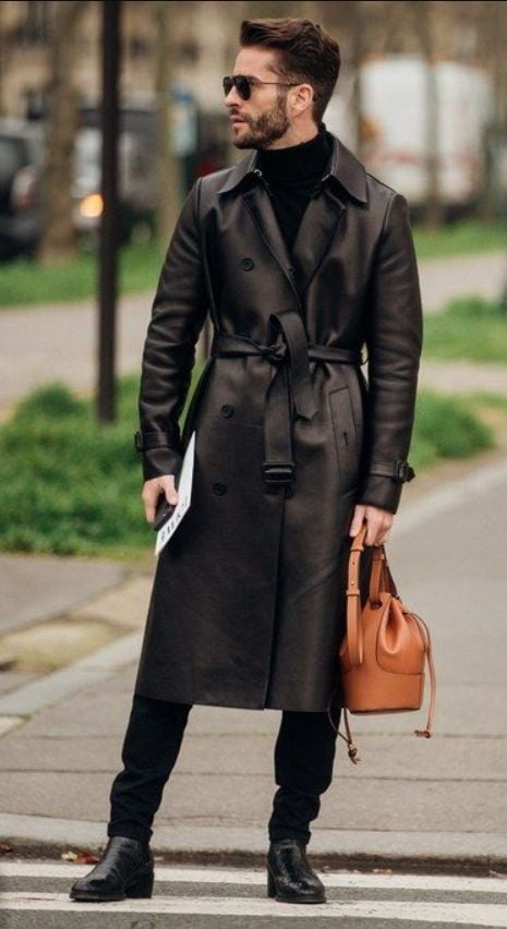 A stylish man in a black leather trench coat, black turtleneck, and sunglasses stands on a city sidewalk holding a tan bucket bag