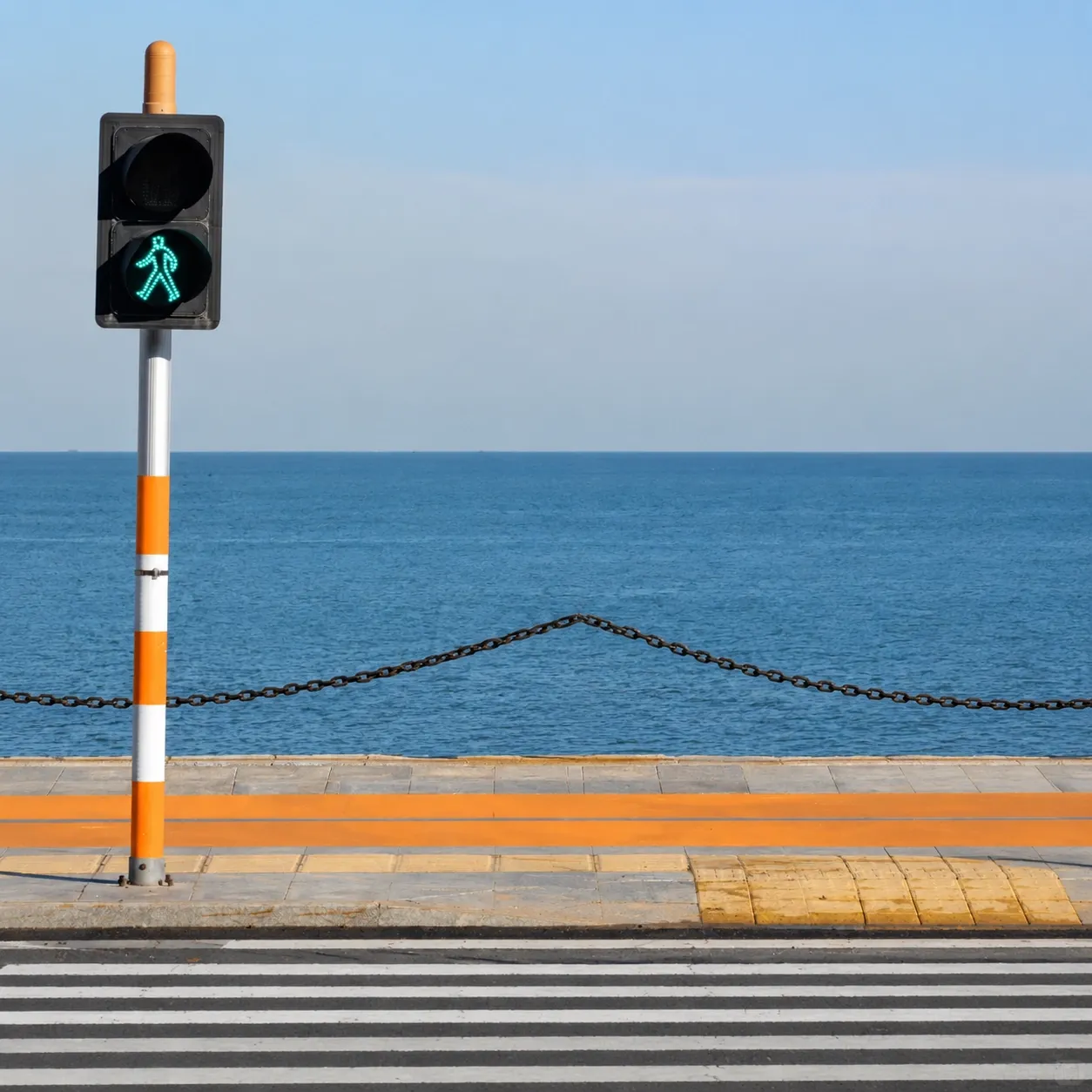 A green pedestrian signal beside a calm sea and a striped crosswalk.