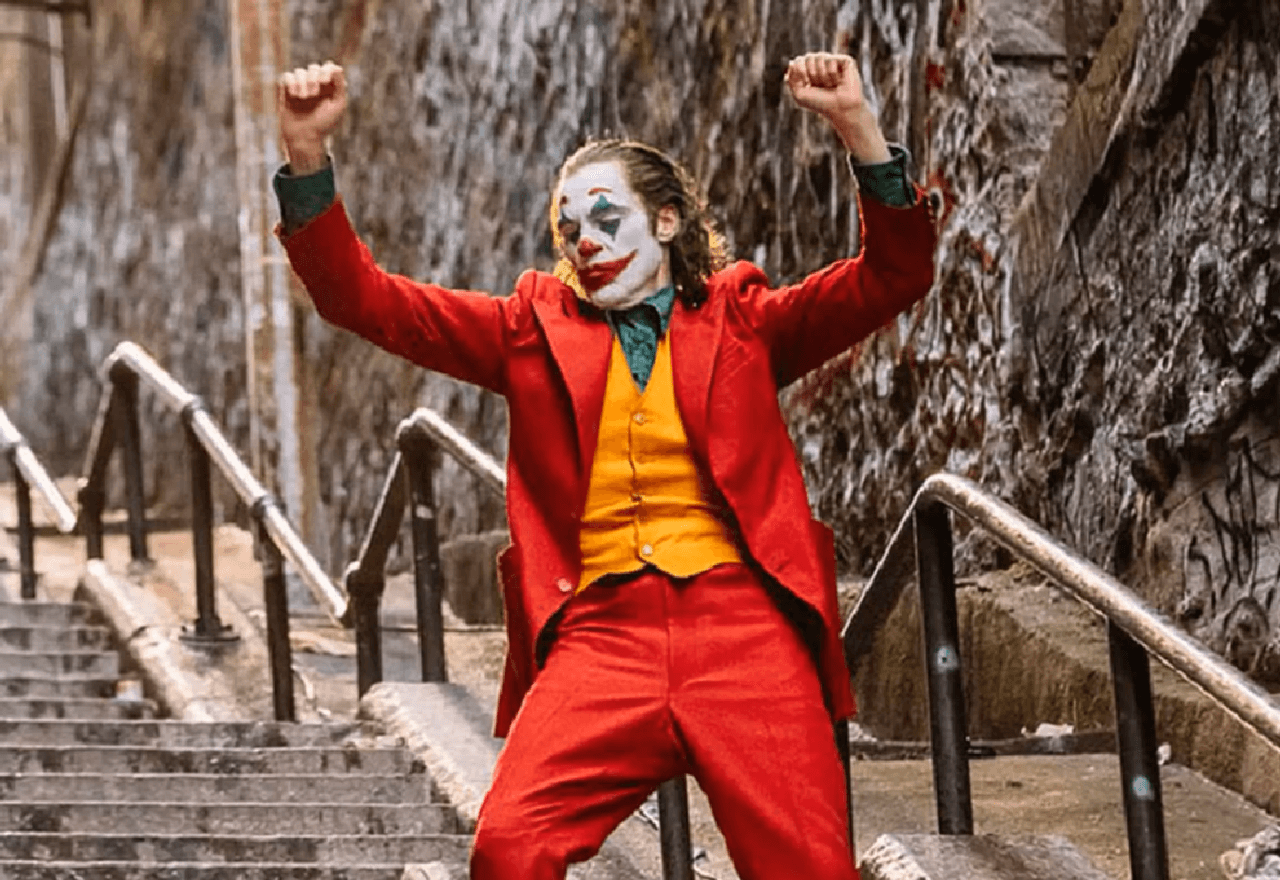 Un payaso con un traje rojo bailando en unas escaleras de la ciudad con los brazos en alto.