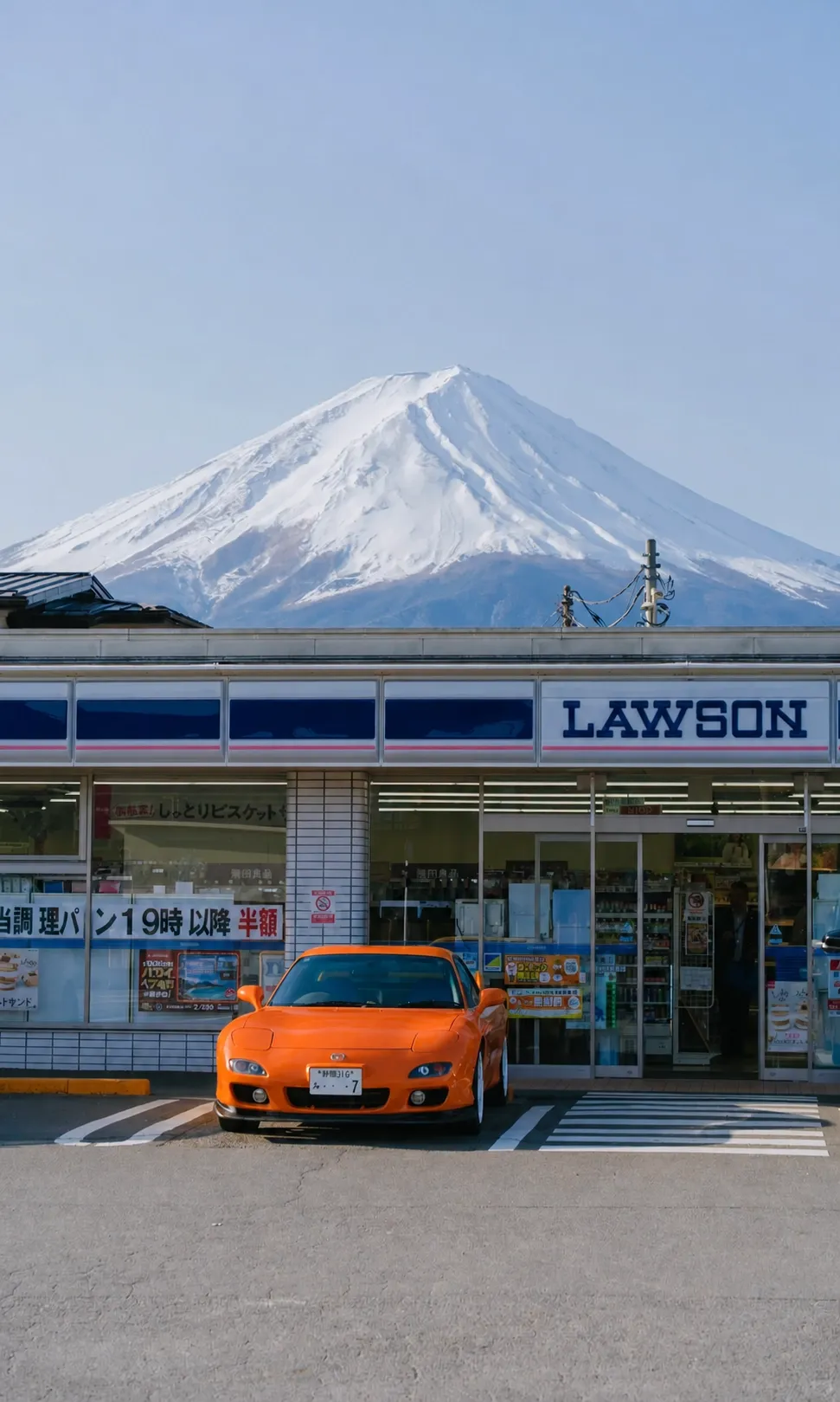 A Lawson convenience store with Mount Fuji in the background and an orange car in front.