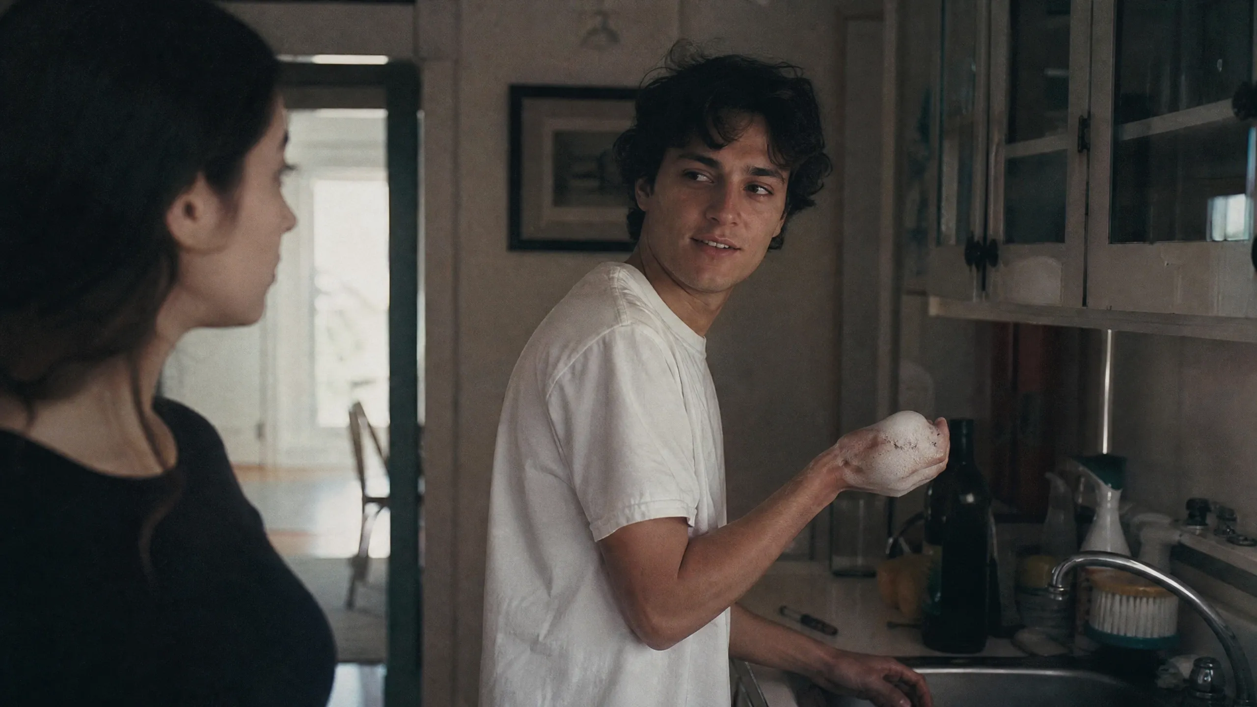 A young man in a white shirt washes dishes while a woman looks on.