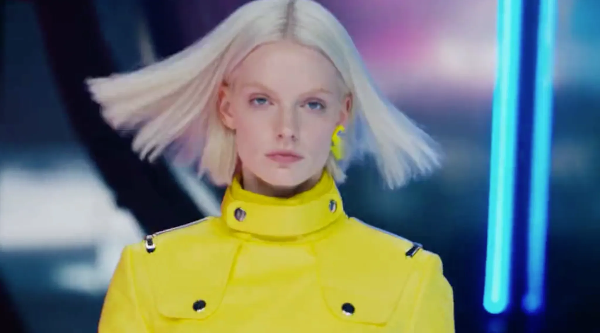 A model with blonde hair wears a bright yellow jacket on a runway.