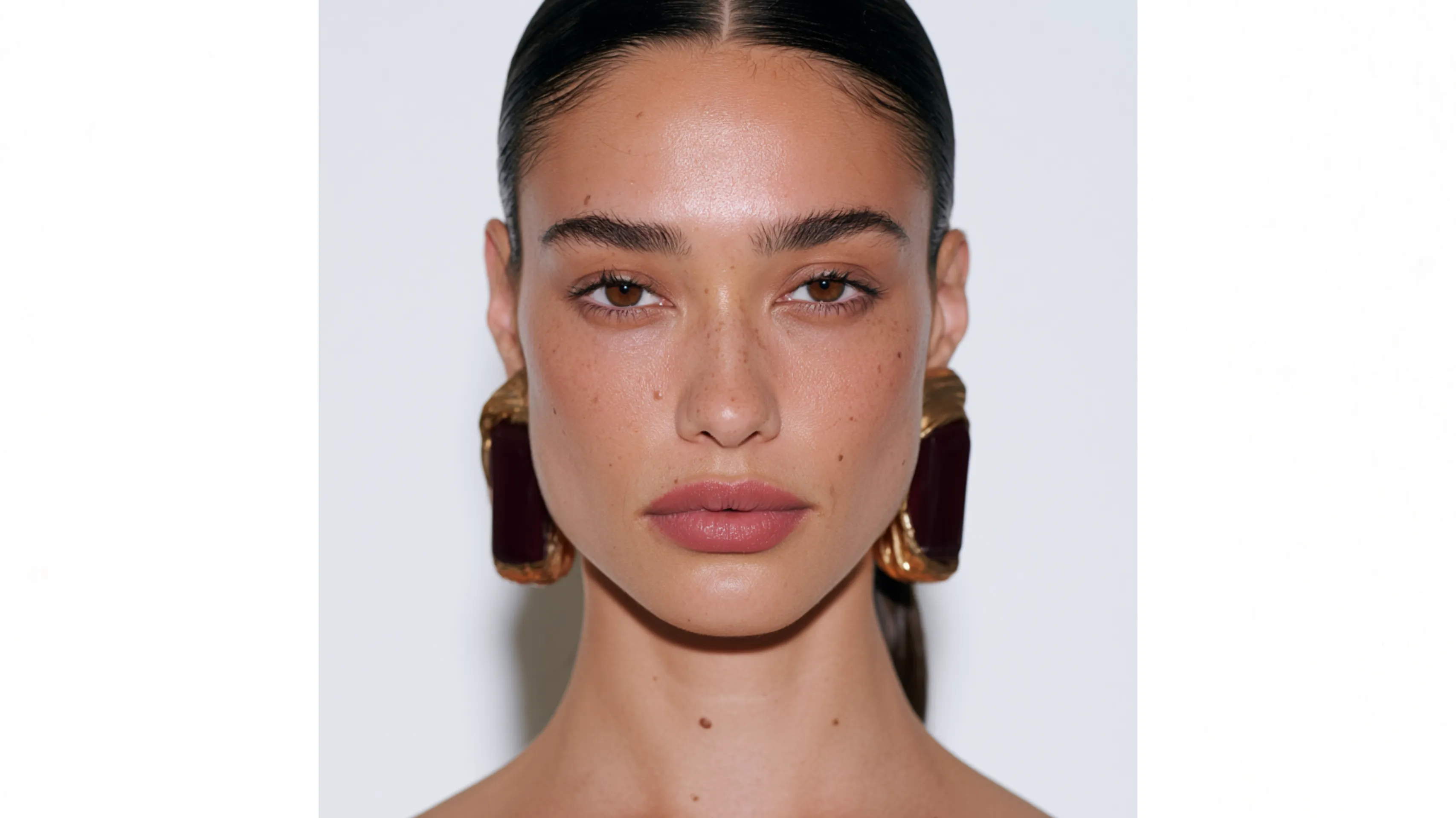 A model with large gold and burgundy earrings poses against a white background.