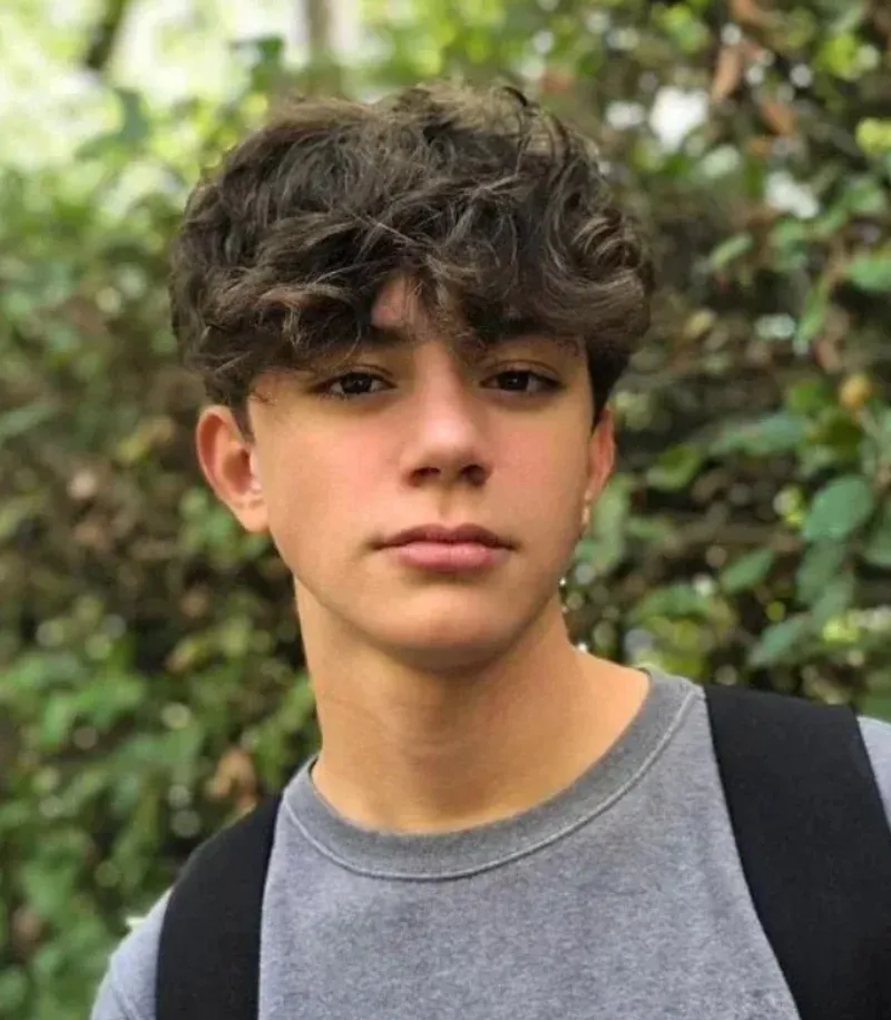 A teenage boy with curly hair stands outdoors in front of greenery.