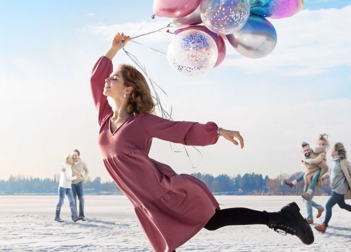 A woman in a pink dress holds balloons while jumping on snow, with a family in the background