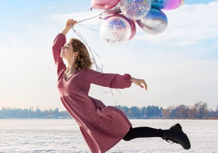 A woman in a pink dress holds balloons while jumping on snow, with no people in the background