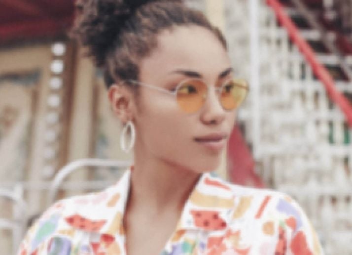 A blurred portrait of a woman with curly hair in a high bun wearing orange-tinted round glasses