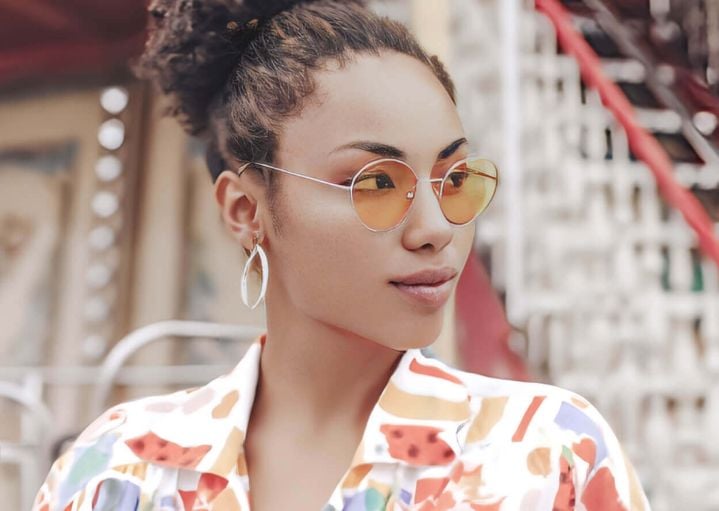 A woman with curly hair in a high bun wears orange-tinted round glasses and a colorful patterned shirt
