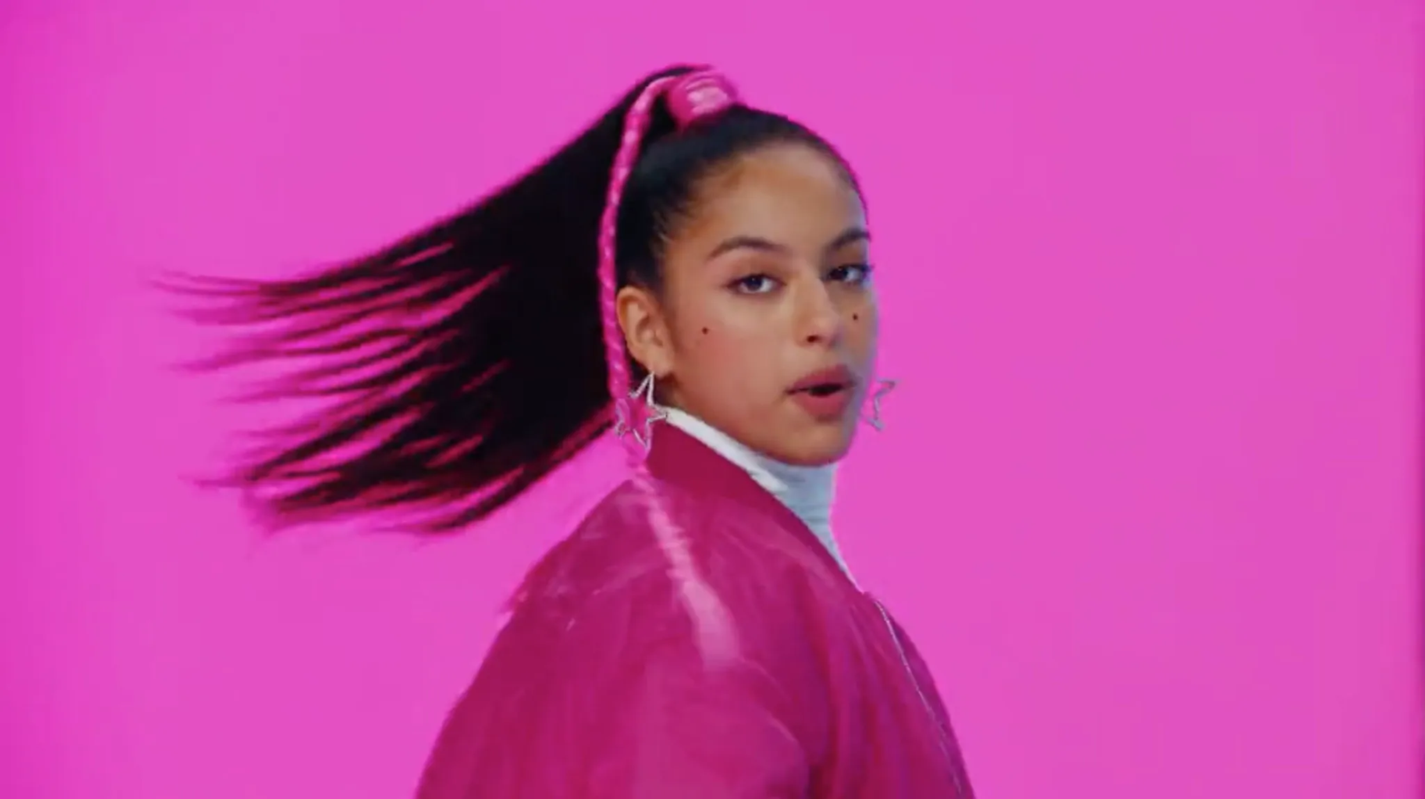 A young woman with long hair turns, wearing a pink jacket and star earrings.